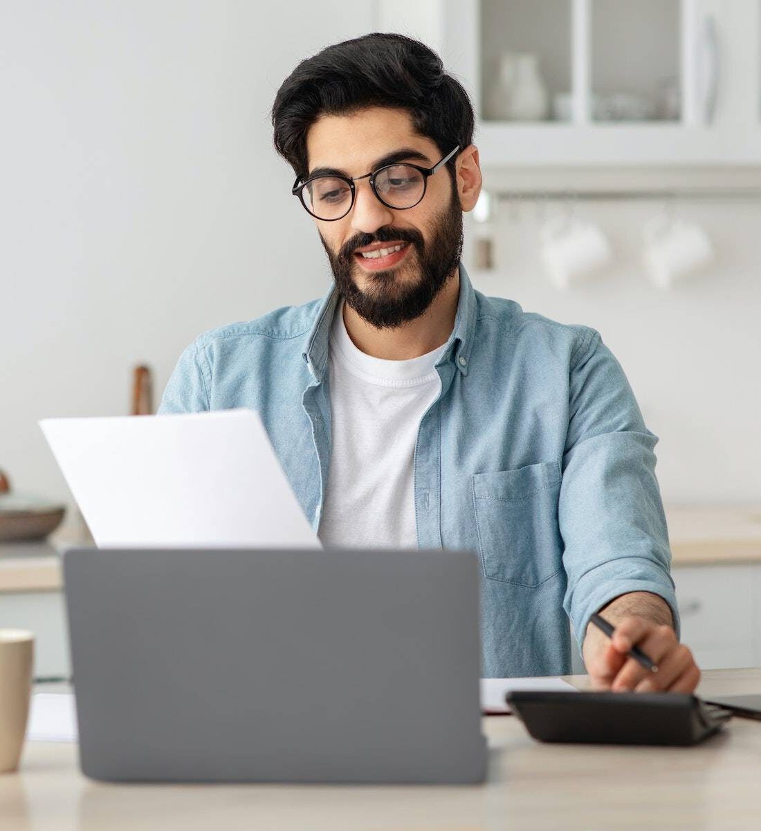 Portrait of happy arab guy reading insurance documents at home, sitting at table in kitchen and